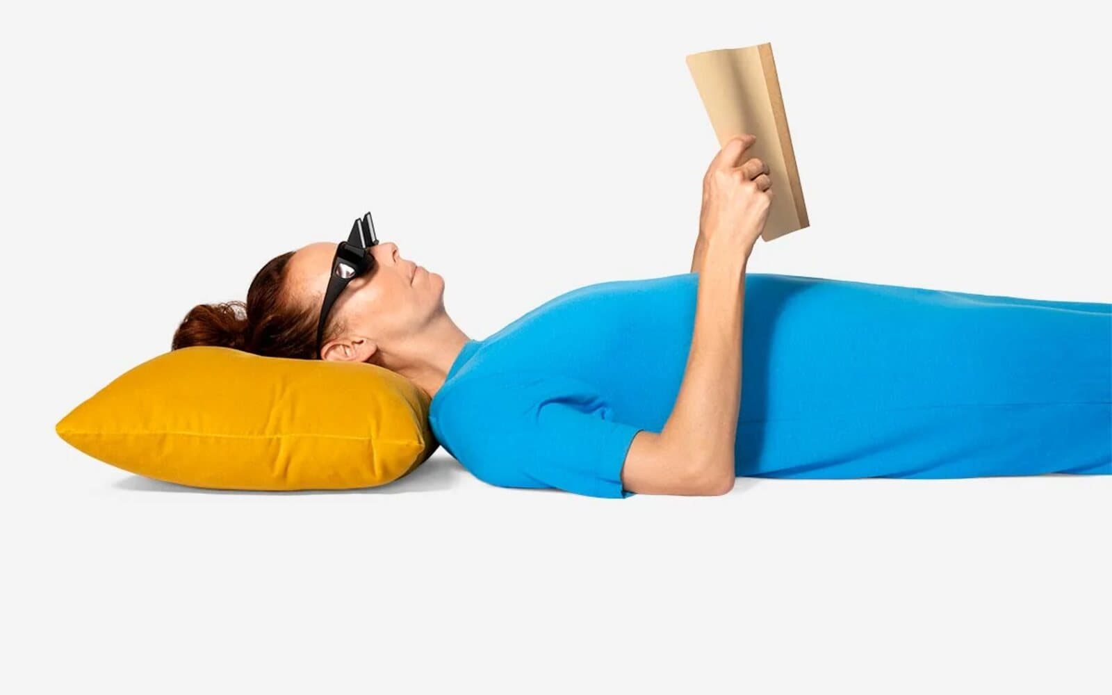 Person lying flat on a pillow reading a book held upright while wearing prism glasses